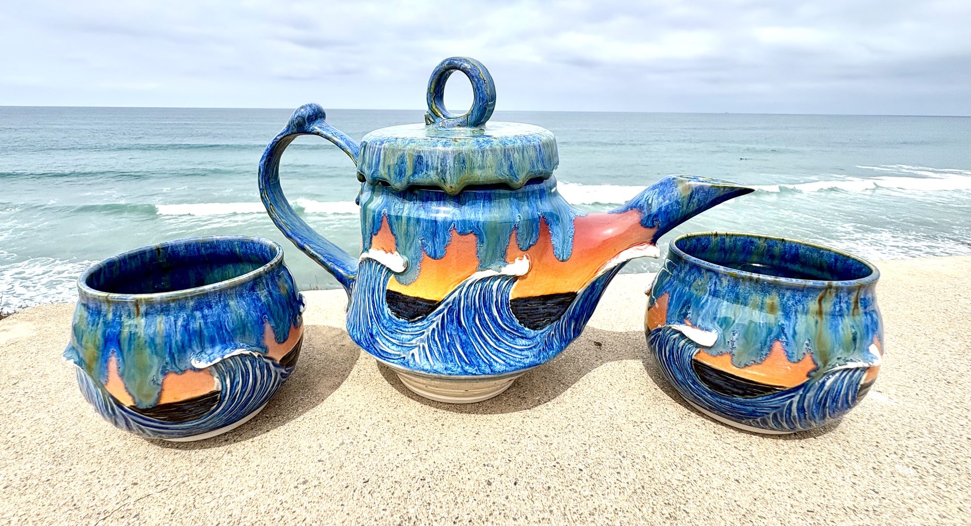 Wave-glaze tea set — teapot and two cups — on a bluff above the surf